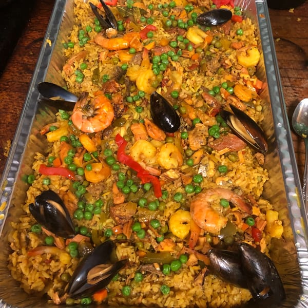 Large foil tray filled with colorful paella, featuring rice, shrimp, mussels, peas, and peppers.