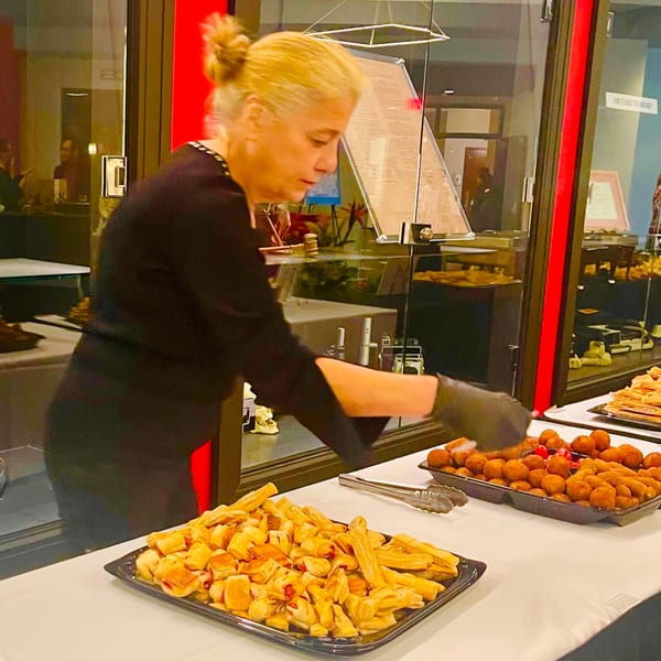 A woman in black gloves arranges pastries and fried snacks on a white-clothed buffet table.