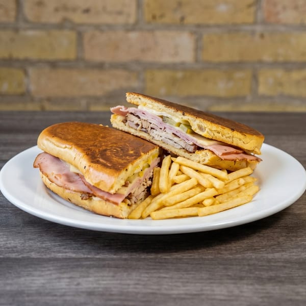 A Cuban sandwich, cut in half, with fries on a white plate against a brick wall.